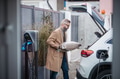 Man putting bag of wood pellets in car trunk, while his electric vehicle is charging. - PhotoDune Item for Sale