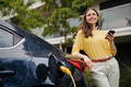 Young woman with smartphone waiting while her electric car charging in home charging station - PhotoDune Item for Sale