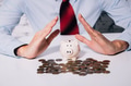 Businessman making hands to protect and save piggy bank of coins and looks like residential house - PhotoDune Item for Sale