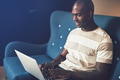 Smiling African entrepreneur sitting on a couch working online - PhotoDune Item for Sale