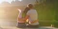 Rear View Of Loving Couple Sitting On Wooden Jetty By Lake At Sunset With Lens Flare - PhotoDune Item for Sale