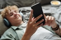 Teenage Caucasian Boy Wearing Headphones Lying on Bed Engrossed in Smartphone - PhotoDune Item for Sale