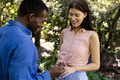 Man proposing to smiling woman in park, holding engagement ring with joy - PhotoDune Item for Sale