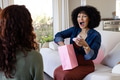 Excited woman opening gift bag on couch, celebrating with friend at home - PhotoDune Item for Sale
