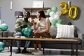 Friends celebrating 30th birthday at home, laughing and holding balloons on sofa - PhotoDune Item for Sale
