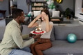 African American man surprising woman with birthday cake, both smiling on sofa - PhotoDune Item for Sale