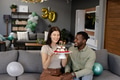 Celebrating birthday at home, couple smiling with cake and candles on sofa - PhotoDune Item for Sale
