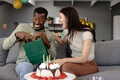 Opening gift bag, African American man and woman laughing, celebrating together on couch - PhotoDune Item for Sale