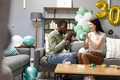 Celebrating birthday, diverse couple inflating balloons in cozy living room setting - PhotoDune Item for Sale