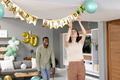 Decorating for birthday party, woman hanging banner while man smiling in living room - PhotoDune Item for Sale