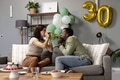 Couple celebrating birthday at home, blowing party horns on couch, feeling joyful - PhotoDune Item for Sale