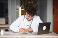 Arab male doctor writing notes on clipboard while working on laptop - PhotoDune Item for Sale