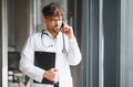 Arab male doctor wearing white coat and stethoscope talking on phone holding clipboard - PhotoDune Item for Sale