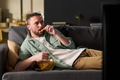 Young Adult Man Reclining on Sofa Eating Chips While Watching Screen - PhotoDune Item for Sale